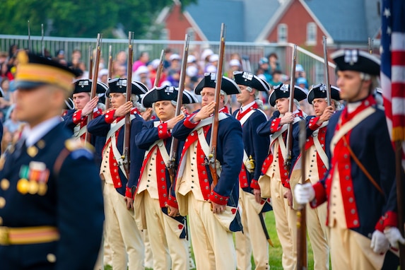 Soldiers wearing Revolutionary War-period uniforms (dark blue with red trim coats, cream-colored pants and dark blue with white trim tri-cornered hats) are adjusting the rifles that they are carrying against their shoulders. In the foreground is a soldier in a modern Army dark ceremonial uniform (dark jacket with gold buttons and dark wheel hat)