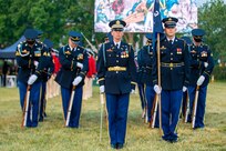 Soldiers in dark ceremonial uniforms (dark jackets with gold buttons, lighter blue pants with gold stripes down the side of the leg, dark wheel hats with rims) have rifles in front of them and are looking down as they affix bayonets to the rifles. The person in the front of the formation has a sword while the person to her left has a guidon flag against his right shoulder.