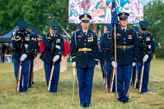 Soldiers in dark ceremonial uniforms (dark jackets with gold buttons, lighter blue pants with gold stripes down the side of the leg, dark wheel hats with rims) have rifles in front of them and are looking down as they affix bayonets to the rifles. The person in the front of the formation has a sword while the person to her left has a guidon flag against his right shoulder.