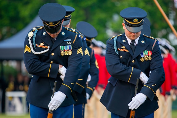 Two Army soldiers are adjusting the bayonets for the rifles, which are propped up against them on the ground, while wearing dark ceremonial uniforms (dark tunics with gold buttons, lighter blue pants, white gloves, dark round caps with bills)