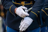 A solder is affixing a bayonet to a rifle. The Soldier is wearing white gloves and a dark ceremonial uniform with shiny metal belt buckle.