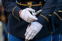 A solder is affixing a bayonet to a rifle. The Soldier is wearing white gloves and a dark ceremonial uniform with shiny metal belt buckle.