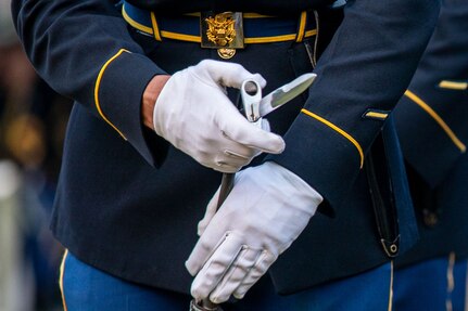 A solder is affixing a bayonet to a rifle. The Soldier is wearing white gloves and a dark ceremonial uniform with shiny metal belt buckle.