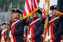 Army soldiers dressed in dark blue with red trim coats from the Revolutionary War period are holding the US Flag and Army flag, and one soldier is holding a period long rifle up against his right shoulder.
