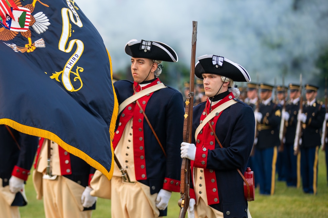 Army soldiers dressed in dark blue with red trim Revolutionary War-era uniforms (and cream colored pants) are in a color guard. One is lowering the U.S. Army flag, and the other is holding a period rifle in front of him. In the background are soldiers dressed in dark ceremonial uniforms holding more modern ceremonial rifles in front of them.