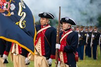 Army soldiers dressed in dark blue with red trim Revolutionary War-era uniforms (and cream colored pants) are in a color guard. One is lowering the U.S. Army flag, and the other is holding a period rifle in front of him. In the background are soldiers dressed in dark ceremonial uniforms holding more modern ceremonial rifles in front of them.