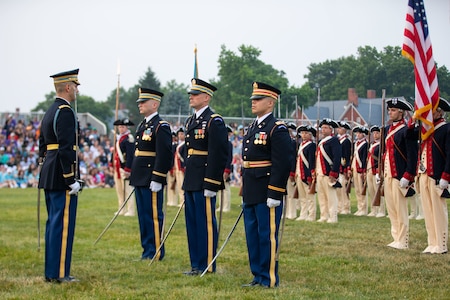 Four Army soldiers in dark ceremonial uniforms are carrying swords at their sides, and they are standing on a green lawn in front of other Soldiers who are carrying the US flag while wearing Revolutionary War-era unifoms (blue coats and white pants).