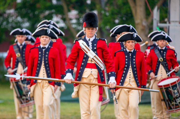 Army soldiers in red Revolutionary War-era uniforms at marching towards the camera. The member in front has a large, dark furry hat on, and is carrying a long stick that looks like a spear. The other members are carrying various instruments.