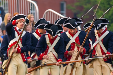 Army Soldiers dressed in Revolutionary War-era blue uniforms with white pants are carrying period rifles with bayonets affixed. They are making adjustments to their rifles.