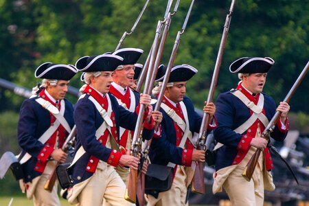 Army Soldiers in dark blue Revolutionary War-era unifoms are running with old style long rifles and baynets. They are wearing tri-cornered hats