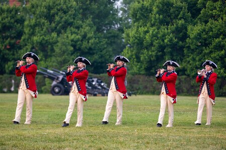 Army Soldiers dressed in red Revolutionary War-era uniforms are marching across a green lawn while playing musical instruments. There are black cannons in the background.
