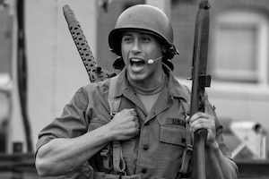 Black and white picture of an Army Soldier dressed in a Korean War-era uniform. He is carrying a rifle in his left hand, and has a machine gun on his back.
