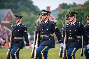 Army soldiers wearing dark ceremonial uniforms are carrying ceremonial rifles with bayonets affived in their right hands while marching