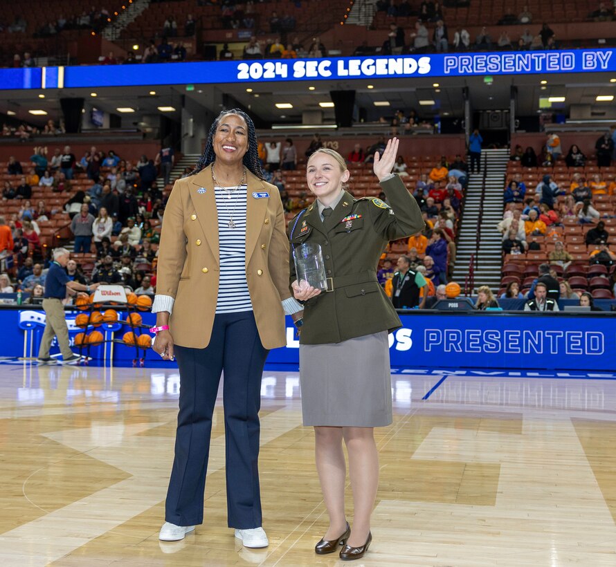 Female Soldier in AGSU poses waving while holding a trophy, standing beside a women in business casual on a basketball court