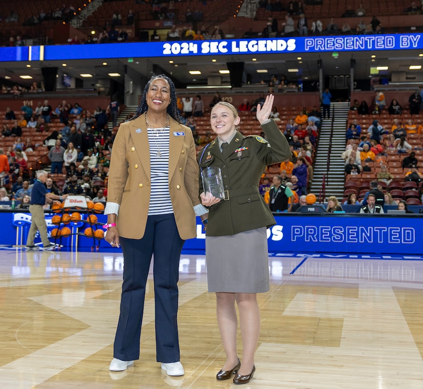 Female Soldier in AGSU poses waving while holding a trophy, standing beside a women in business casual on a basketball court