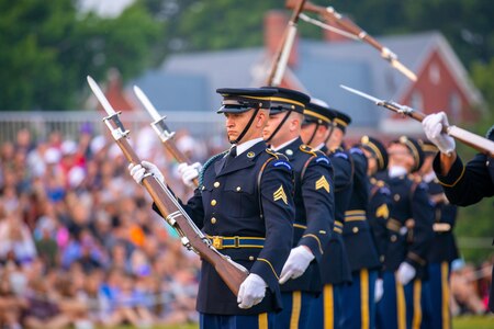 Army soldiers in dark ceremonial uniforms are spinning ceremonial rifles with bayonets affixed in front of them. They are standing in row. In the background, the audience is sitting on bleachers.