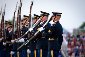 Army soldiers in dark ceremonial uniforms are spinning ceremonial rifles with bayonets affixed in front of them. They are standing in row.