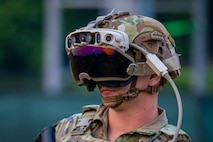 Army soldier in Army Combat Uniform (green camouflage) is wearing a helmet with large dark goggles that has a cable attached to it.