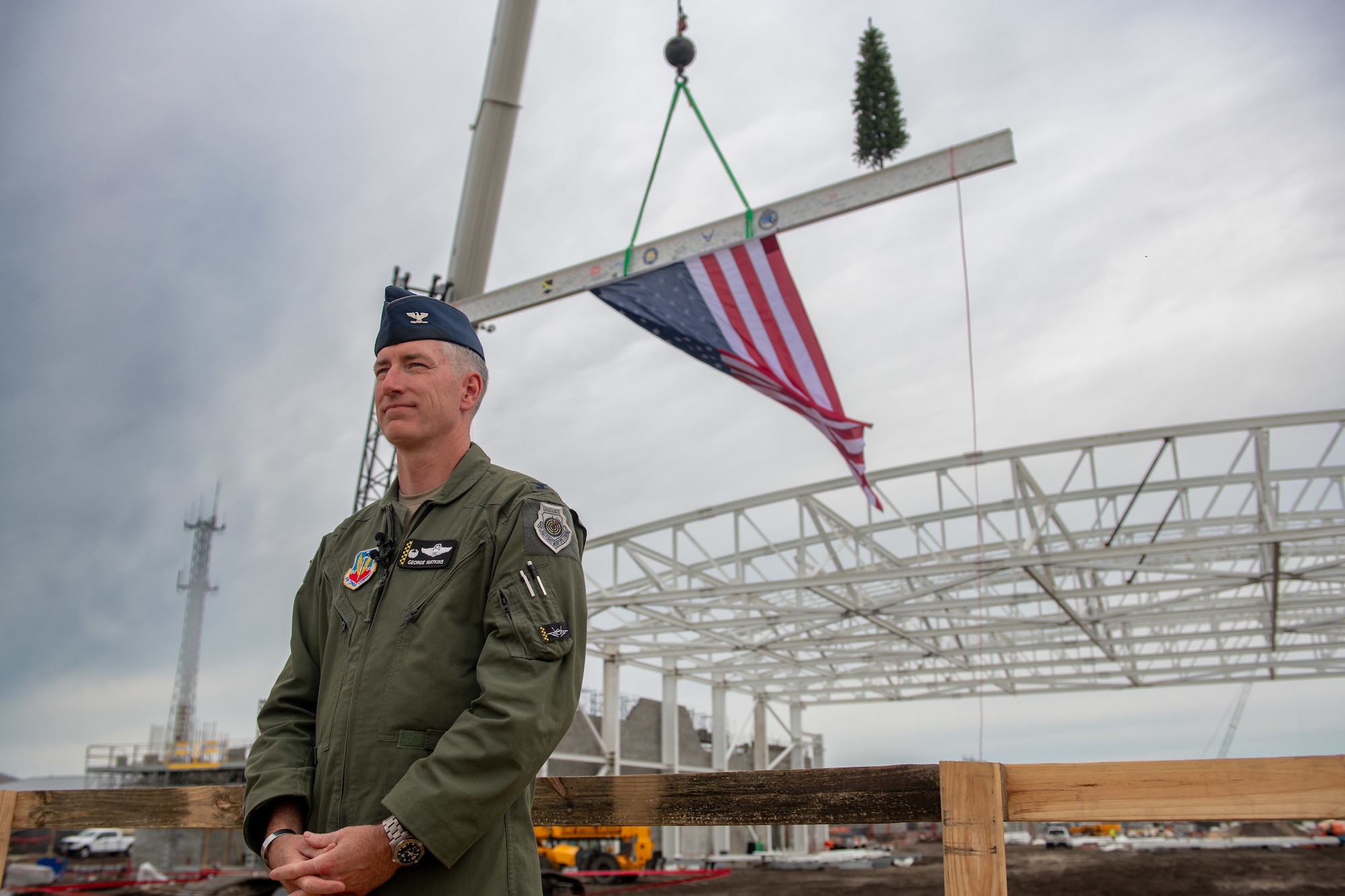 Man in military uniform stands in front of American flag on a construction site.