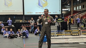 A uniformed service member holds a microphone and speaks as young people seated on the floor look up.