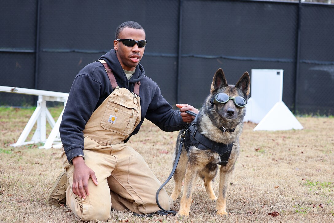 A soldier kneels alongside a military working dog while holding its leash. Both are wearing sunglasses.