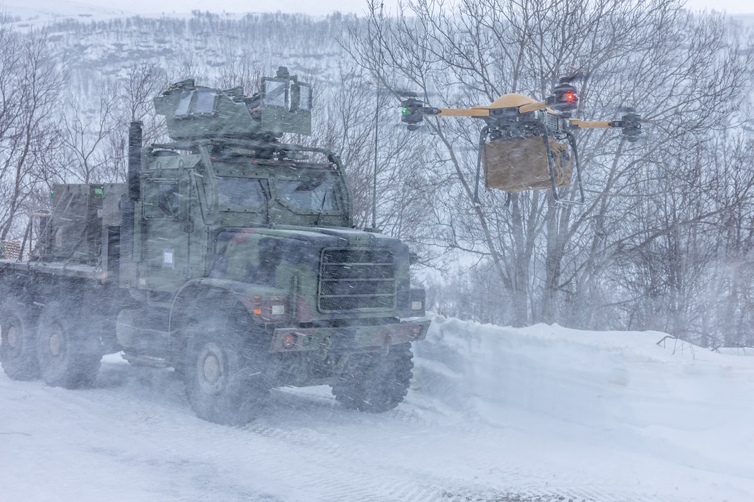 An unmanned aerial system hovers in front of a military vehicle in snowy weather.