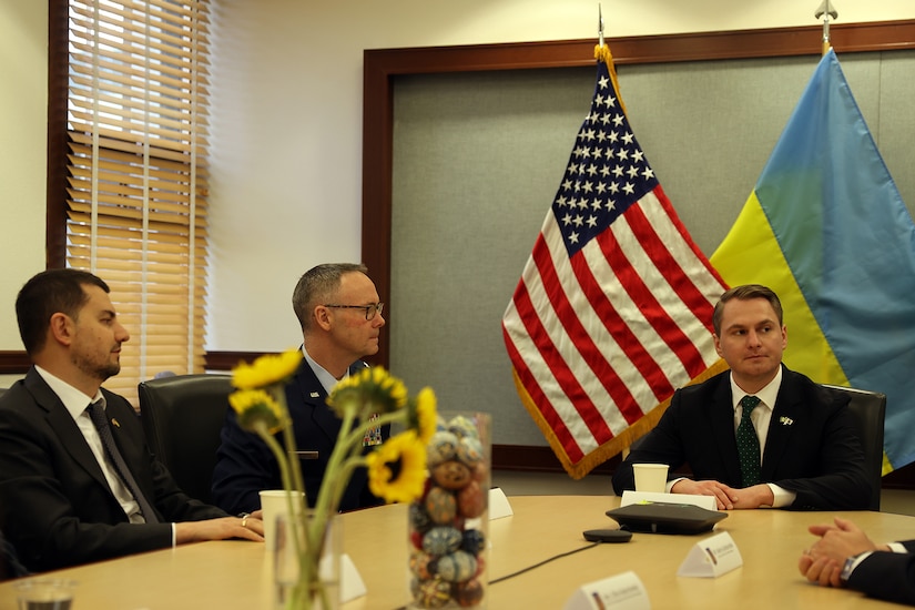 Three people sit at a table with flags in the background.