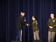 man wearing u.s. army uniform shakes the hand of a women wearing u.s. army uniform on a stage.