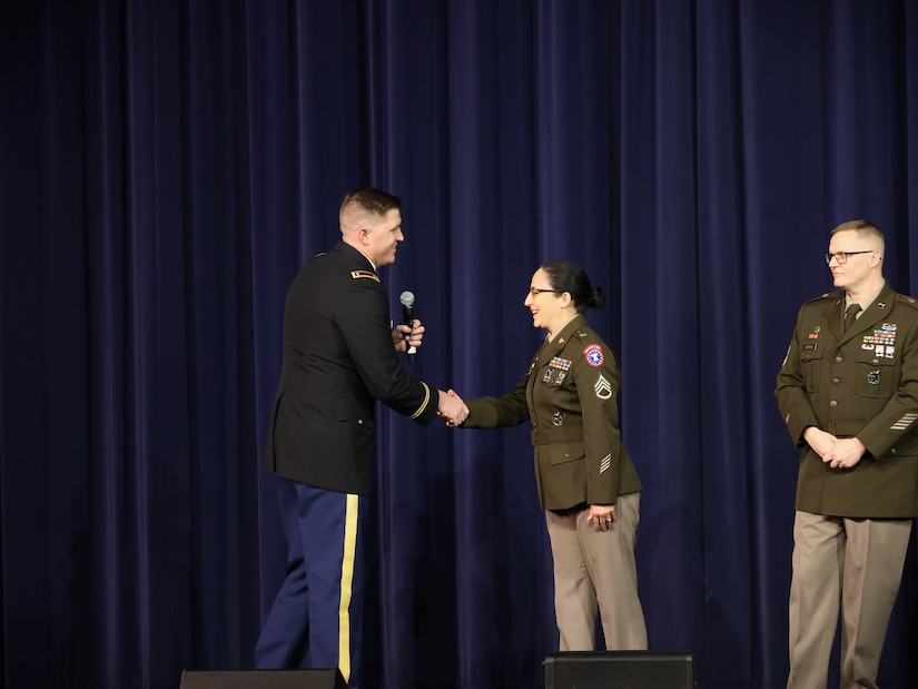 man wearing u.s. army uniform shakes the hand of a women wearing u.s. army uniform on a stage.