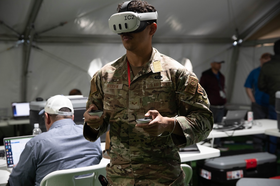 An airman wearing a virtual reality headset holds a control device in each hand while standing in a white tent with other people on computers.