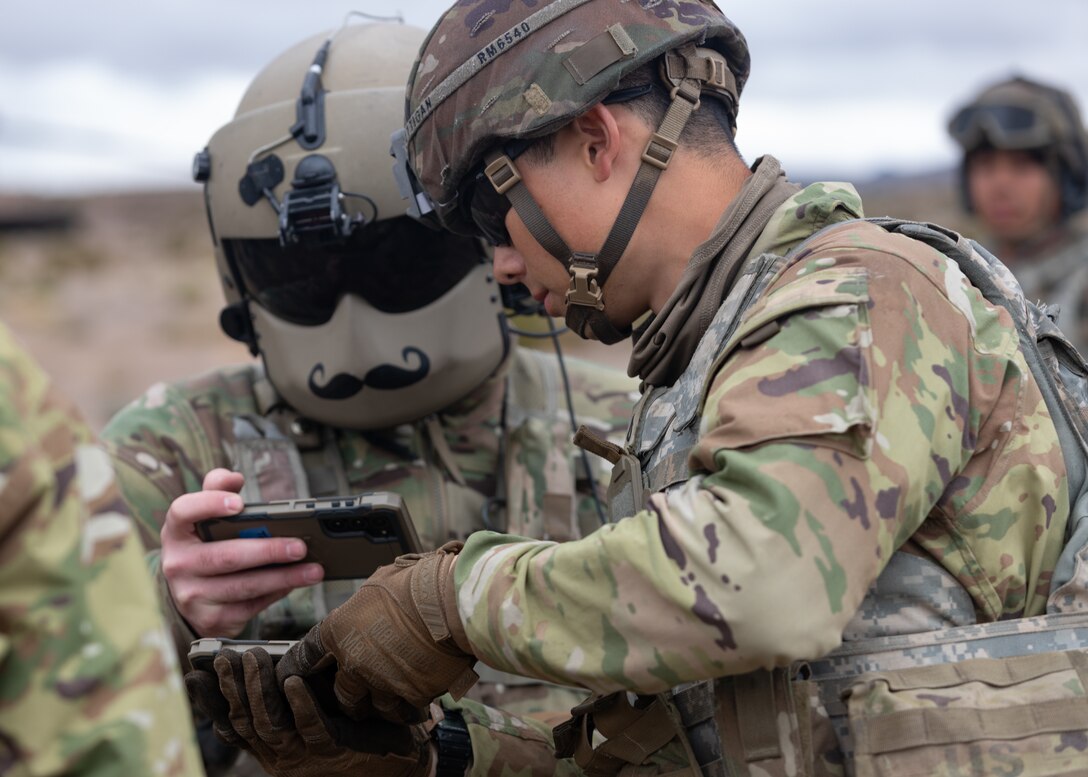 A soldier wearing a helmet and mask with a handlebar mustache holds a smartphone for another soldier to view with one soldier in front of them and one behind them.