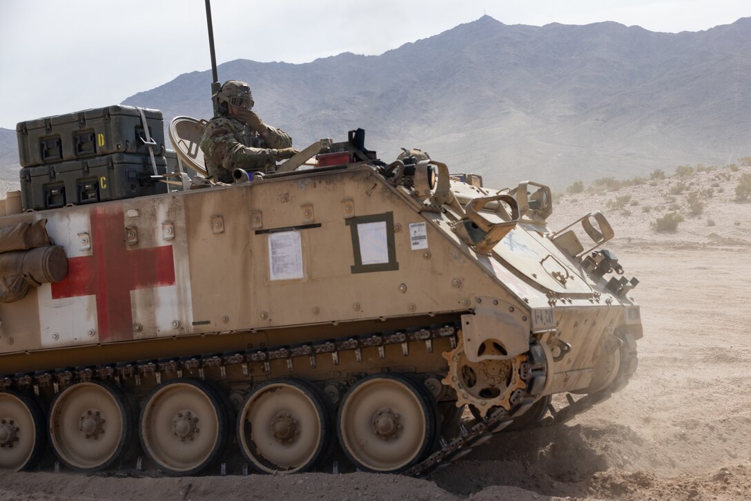 A tank with a first aid symbol moves through a desert area with a soldier driving on top.