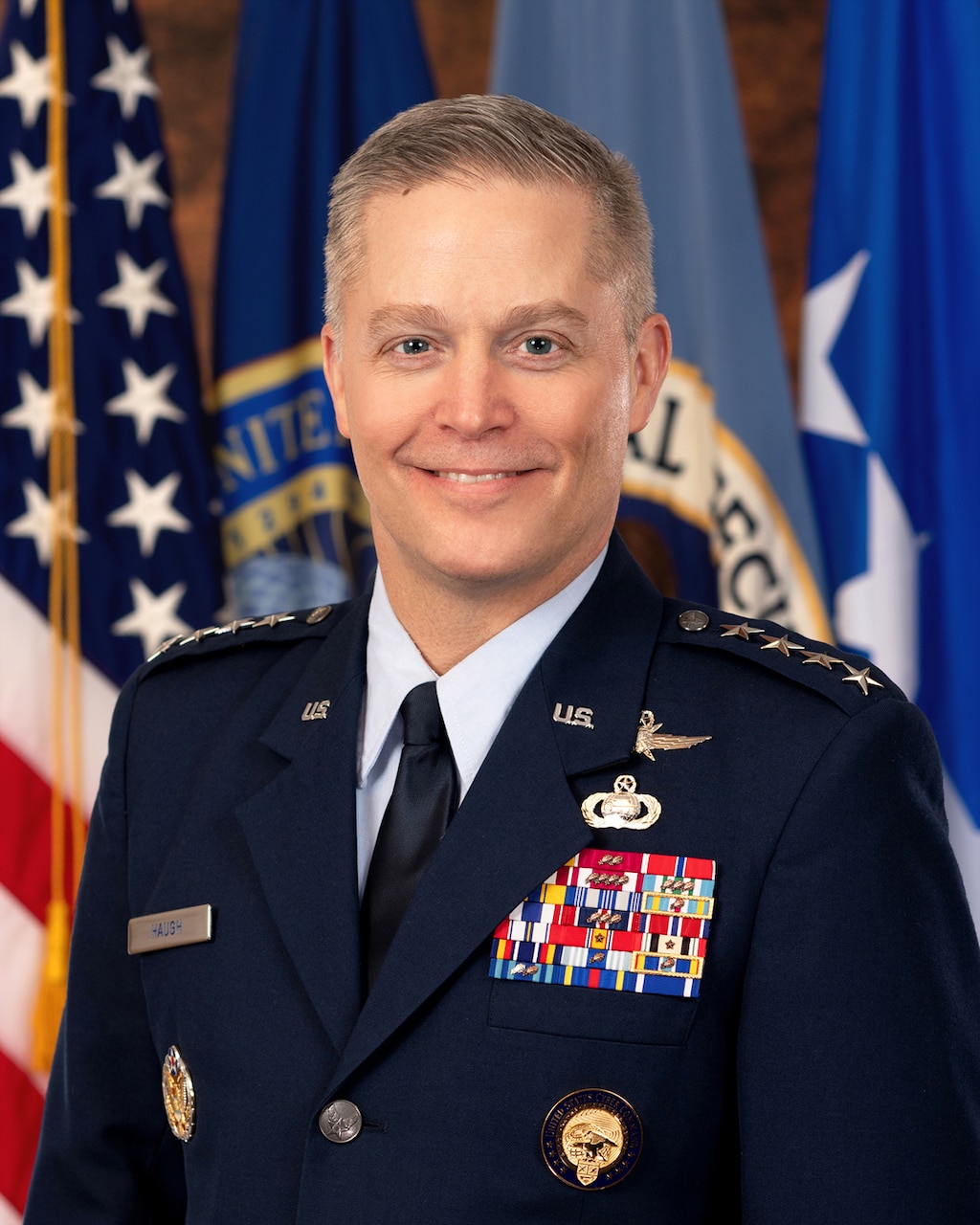 A white man with blonde hair in a U.S. Air Force uniform poses facing the camera in front of several flags.