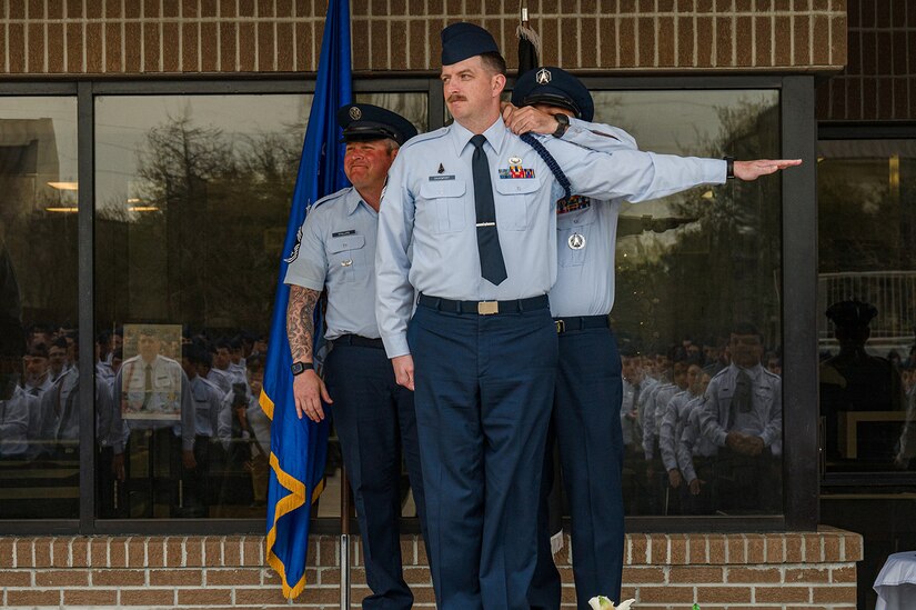 A Space Force member stands with their left arm extended while another service member applies a blue braid to the person's shoulder.