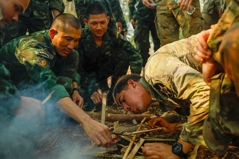 A U.S. soldier blows on a human-made jungle fire as fellow U.S. and Thai soldiers huddle around it.