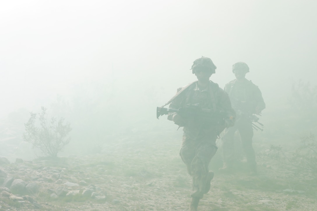 Two soldiers in tactical gear carrying weapons walk through a hazy, desert-like area.