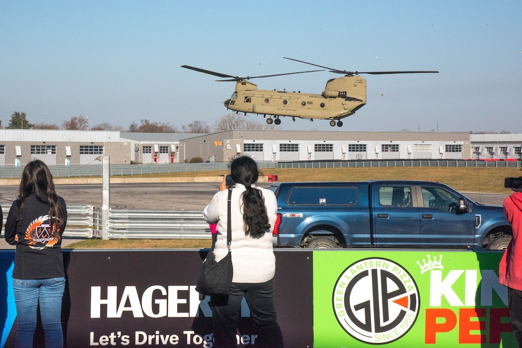 Dual rotor CH-47 tan-colored helicopter hovers over field. Students watching.