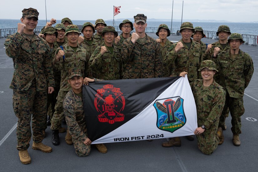 U.S. Marines and Japanese soldiers pose for a photo holding the Iron Fist 24 flag with water and the rising sun flag in the background.