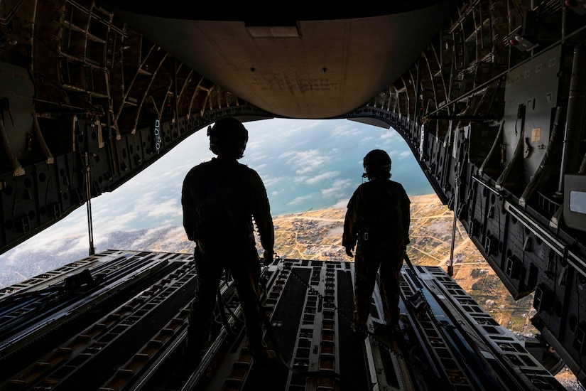 Two airmen stand and look out from the open ramp of an airborne aircraft with clouds and land in the backdrop.