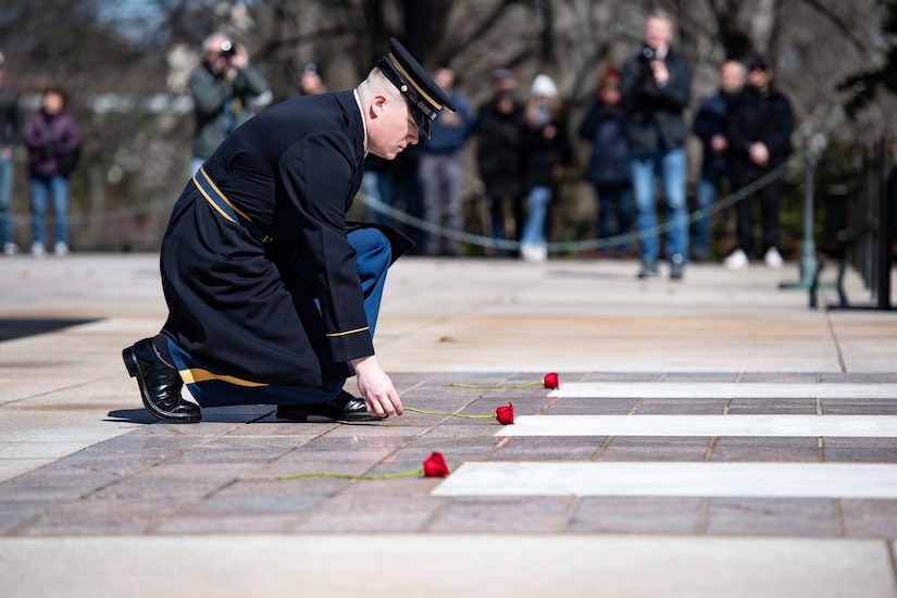 A soldier in a formal uniform kneels down and places three roses on the ground in front of a tomb.