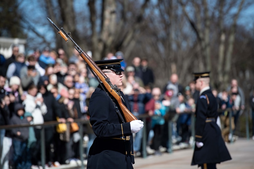 A soldier in a formal uniform holds a rifle and stands at attention in front of a crowd.