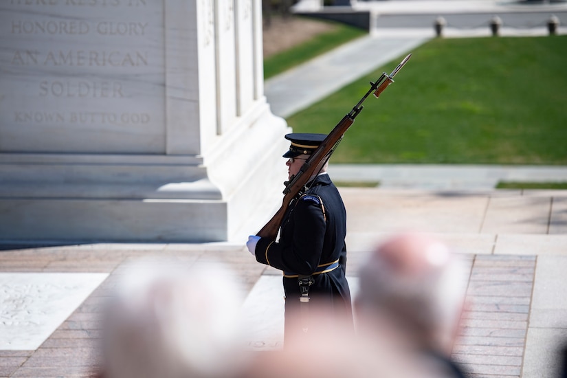 People watch a soldier in a formal uniform as he walks in front of a tomb.