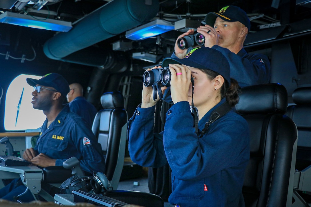 Three sailors, two holding binoculars, look forward. Two of the sailors are seated.