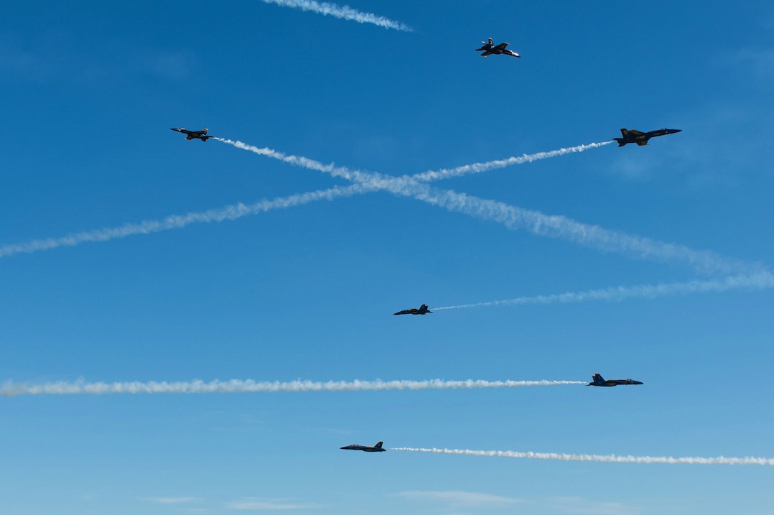 Military jets leave white trails of smoke as they fly in all directions across a blue sky.