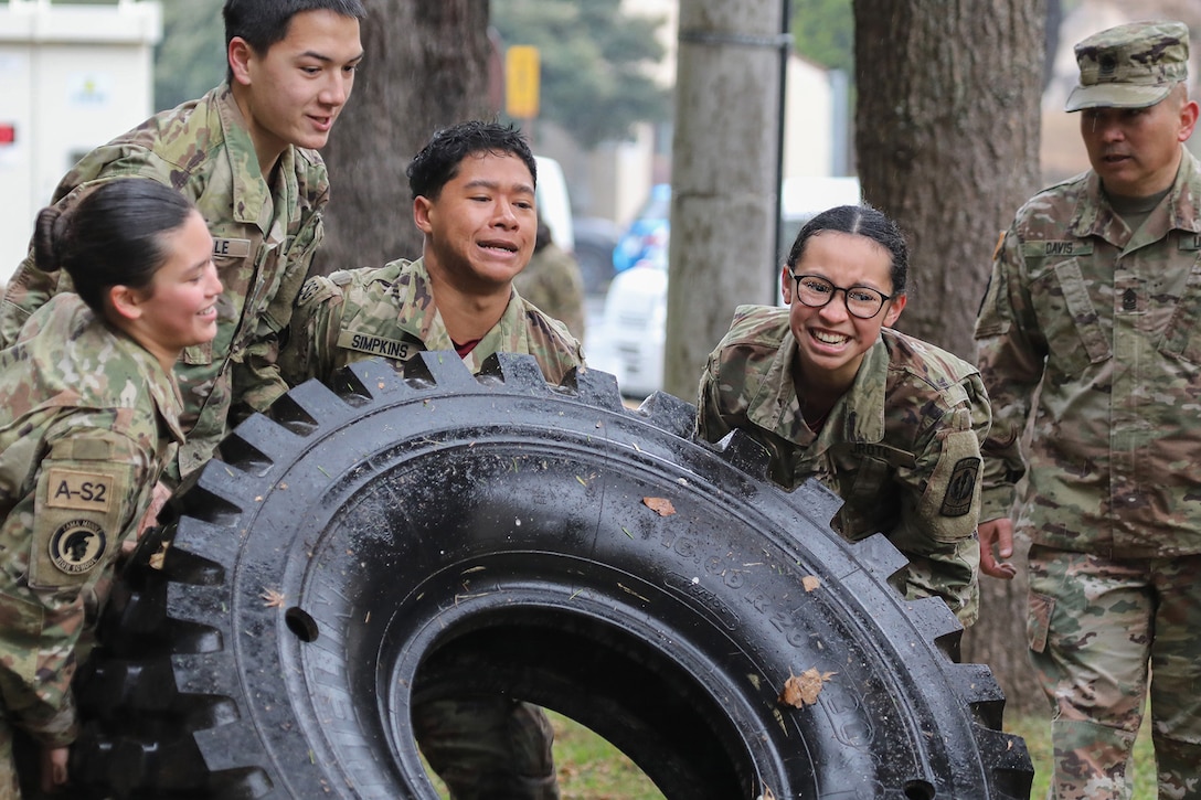 Four junior ROTC cadets work to flip a tire as a soldier watches.