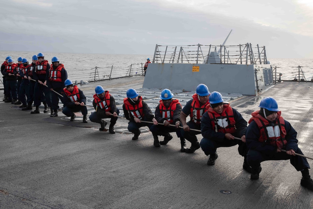 Fifteen sailors wearing helmets pull a rope line on the flight deck of a ship at sea. Seven of the sailors are in a squatting position.