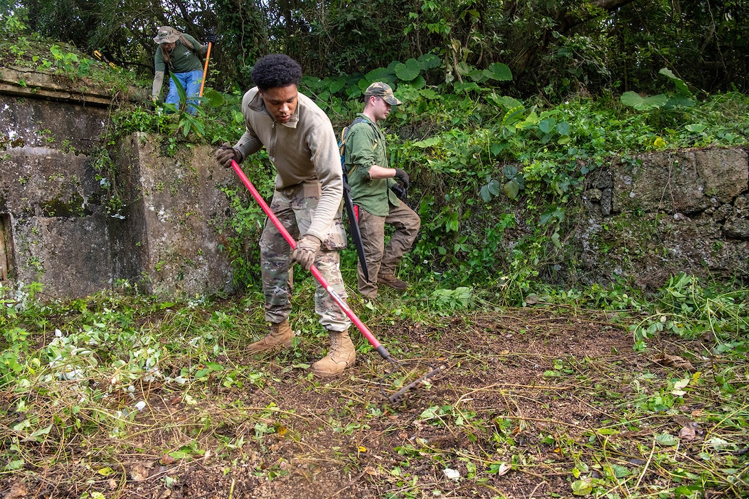 An airman uses a rake to clear vines and grass from an area as two people work in the background.