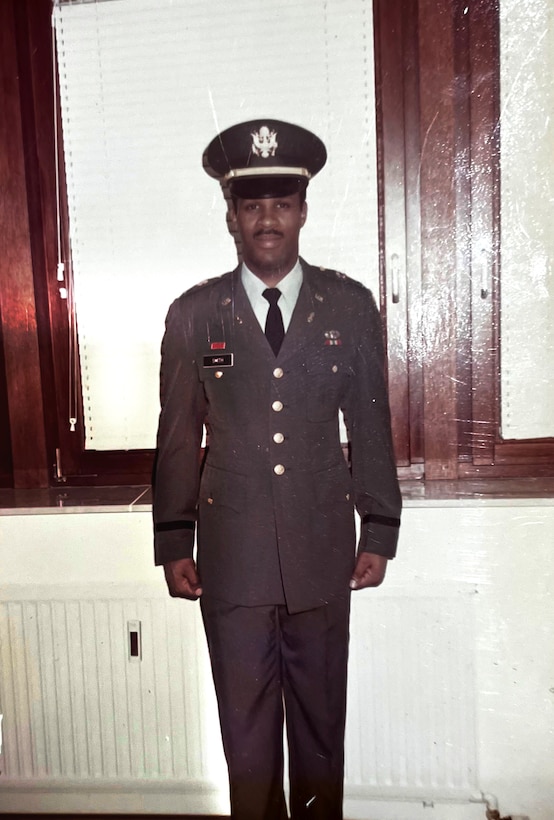 Black man stands at attention indoors in an Army dress green uniform.