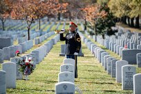 an Army soldier wearing a dark ceremonial uniform (dark tunic top with gold buttons, blue pants, bright red wheel hat with rim) is playing a trumpet in a cemetery with rows of white rounded marble tombstones