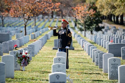 an Army soldier wearing a dark ceremonial uniform (dark tunic top with gold buttons, blue pants, bright red wheel hat with rim) is playing a trumpet in a cemetery with rows of white rounded marble tombstones